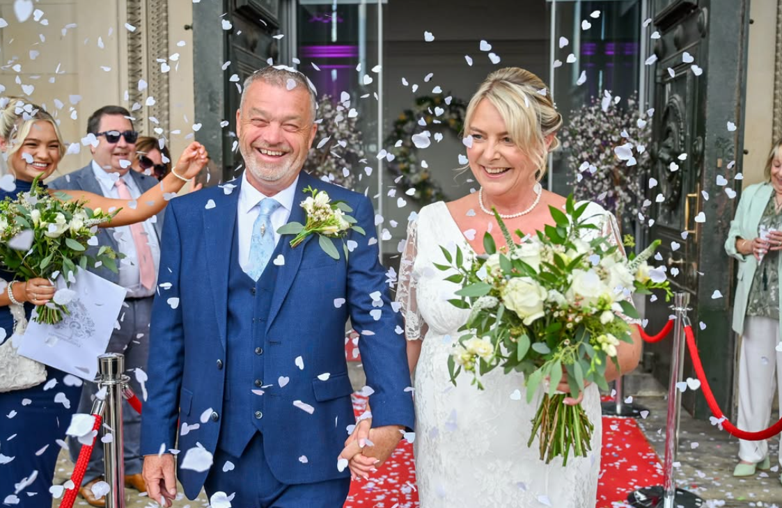 Newlywed couple laughing as guests throw confetti outside a wedding venue